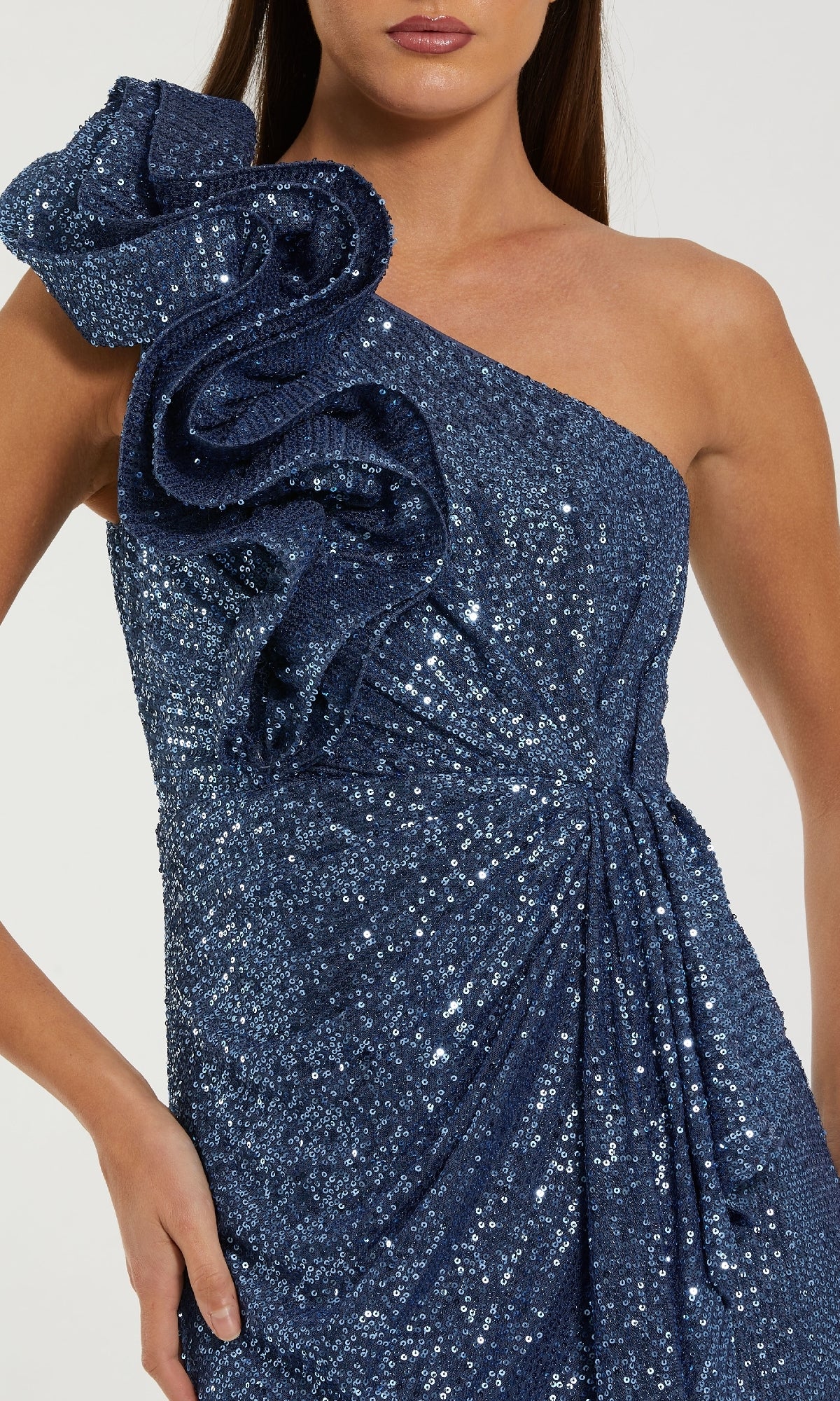 A woman stands confidently with one hand on her hip, wearing the Mac Duggal 50093—a long blue sequin formal gown featuring a striking one-shoulder design with a bold ruffled shoulder detail.
